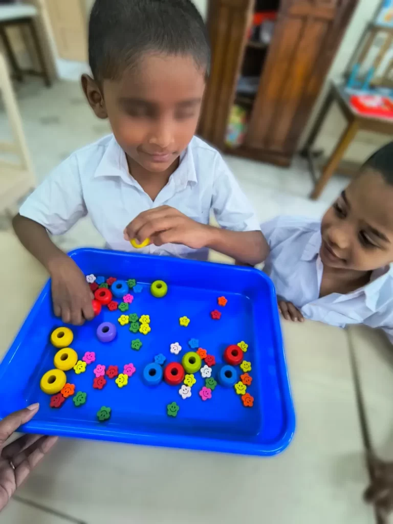In the picture, a child with visual impairment is feeling out various shaped beads in a tray extended to him. His friend watches on.