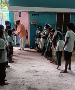 This image is of a teacher from Vellore School, who is distributing chocolates on Children’s Day on behalf of VE, while the kids are standing in parallel lines facing each other