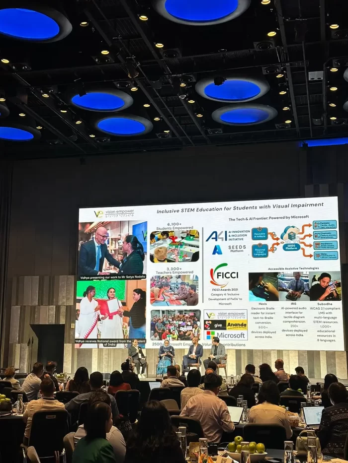 Vidhya speaking to a panel of three people on stage. Behind the stage, there is a large screen, showcasing various awards and projects of VE.