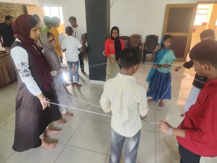 Students and participants at the workshop taking part in the computational thinking games, where they are all standing together holding a thread connecting through a circle